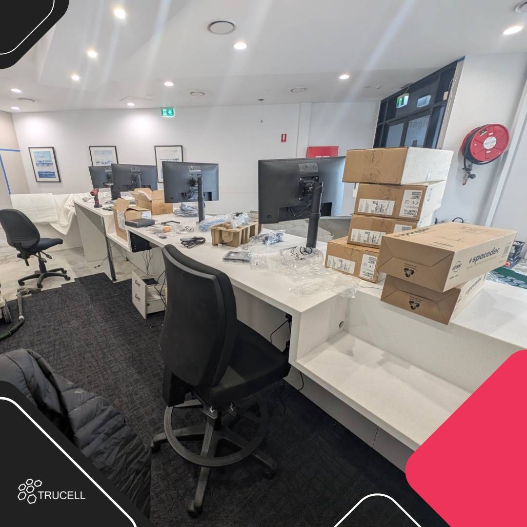 Reception workstations partially installed at Medical Imaging Erina with dual LG monitors on adjustable arms, hardware boxes from HP and Spacedec staged ready for deployment, drawer pedestals in position, and CPU mounts cabled under the bench